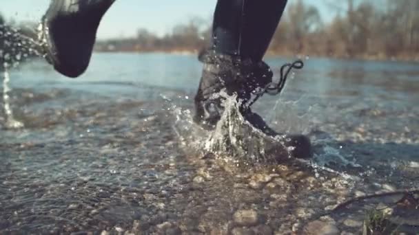 慢动作 低角度 在一个美丽的秋天 穿着黑色防水靴的女孩在清澈的湖水中飞溅 同时在浅水处跑来跑去 晶莹的水滴飘扬 — 图库视频影像