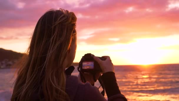 慢动作 年轻的女摄影师在海面上捕捉到美丽的夏夜太阳 轻柔的微风吹拂着女孩的头发 她拍下了落日美景的照片 — 图库视频影像