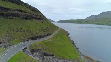 Hava, Kopya Uzay: Araba Faroe Adaları 'ndaki kıyı yolu boyunca ve balık çiftliklerinin yanından geçiyor. Uçsuz bucaksız çimenli tepe ve güzel mavi körfez manzaralı kırsal bölgede turistik araçları çevrelemektedir..