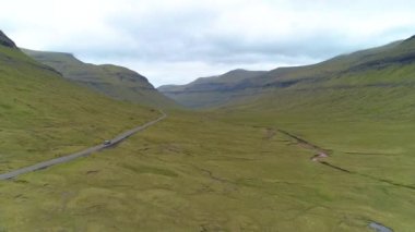 Faroe Adaları 'nın uzak bölgelerindeki manzaralı yeşil bir vadiden geçen dar bir yolda araba sürüyor. Sarp tepeler, bulutlu bir günde turist arabasının etrafını sarar. Düz yolda ilerler..