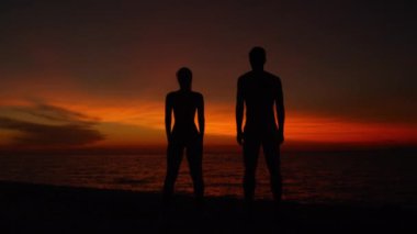Yakından, Silhouette: zinde çift kollarını açarken turuncu günbatımına ve sakin okyanusa bakar. Erkek arkadaş ve kız arkadaş muhteşem renkli bir yaz sabahını kutluyorlar..