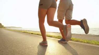 Slow Motion, Low Angle, Lens Flare: Atletik genç kadın ve erkek boş manzaralı bir kıyı yolunda koşuyorlar. Güneşli yaz gününde deniz kenarında antrenman yapan iki tanınmayan fit insanın sinematik çekimi..