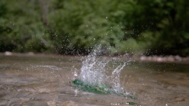 Slow Motion, Close Up: Boş yeşil cam şişe gökten nehre düşüyor. Dağlarda güneşli bir günde saf kristal berrak derede yüzen çöpler. Çöpler doğayı kirletir.