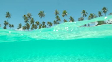 Slow Motion, Half Underwater: kristal berrak okyanus dalgaları Tayland 'da nefes kesici kumlu sahile yaklaşıyor. Parlak yaz güneşi tropikal beyaz kumsal, yeşil palmiye ağaçları ve turkuaz egzotik deniz üzerinde parlıyor..