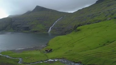 Büyüleyici yeşil kırsal kesimdeki büyük gölün yakınındaki yalnız evin üzerinde uçuyor. Dik tepeler ve otlar Faroe Adaları 'nın kırsal kesimindeki evin etrafını sardı. Nehirler göle akıyor..