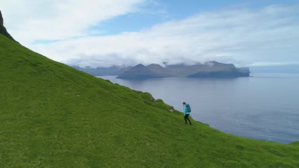 年轻女子沿着长满青草的小山漫步 尽收眼底 环视斯堪的纳维亚海滨 长满青草的大陆和宁静的深蓝色海洋 在法罗群岛度假的女远足者 — 图库视频影像