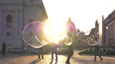 Vatikan, Eylül Slow Motion, Close Up, Lens Flare, Dof: Havalı sokak sanatçıları büyük sabun köpükleri yaparken çocuklar arkalarından koşup onları patlatıyor. Gün batımında yüzen renkli kabarcıklar