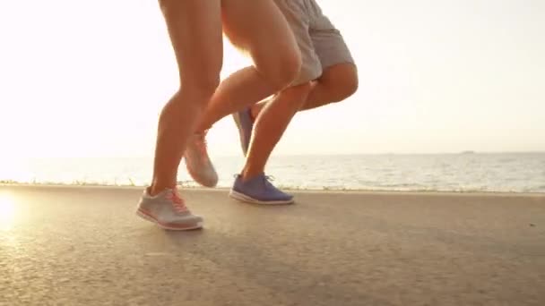 スローモーション 低角度 スロベニアの風光明媚な海岸沿いの道路に沿って実行されている2つのトレーニングパートナー 夏休みにジョギングをする人の後ろに美しい海辺 男性と女性のトレーニングを一緒に — ストック動画
