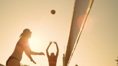 Slow Motion, Low Angle, Close Up, Lens Flare: Bikinili atletik beyaz kadın topu zıplayan adamı geçti ve voleybolda bir puan aldı. Gün batımında plaj voleybolu oynayan formda insanlar..