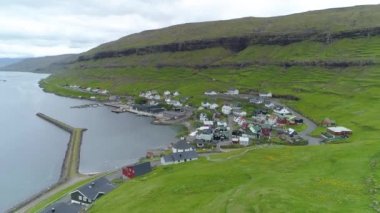 Faroe Adaları 'ndaki sakin sahil kasabasından uçarak canlı otlak tepelerin altına gizlenmiş. Kayalık İskandinav kırsalında bulutlu bir günde muhteşem bir balıkçı köyü manzarası.