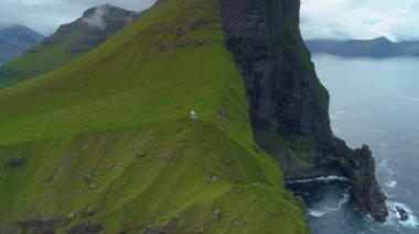 Manzaralı siyah uçurumların ve çimenli adadaki küçük beyaz deniz fenerinin yanında uçmak. Bilinmeyen yürüyüşçüler sonsuz okyanus manzarası ile muhteşem yeşil dağ manzarası etrafında yürürler..