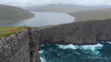 Hava Kopyalama Uzayı: Resimli Faroe Adaları 'ndaki gri bulutlu bir günde yükselen uçurumları ve durgun bir gölü gözlemleyen kadın yürüyüşçü. Nefes kesici engebeli doğa yürüyüşü yapan genç bir kadının muhteşem manzarası.