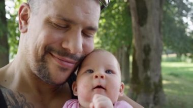 Slow Motion, Close Up, Copy Space: Büyük dövmeli baba yeşil parktaki küçük kızıyla paha biçilmez güzel anlar paylaşıyor. Neşeli, kaslı baba yeni doğmuş güzel kızını yanağından öpüyor..