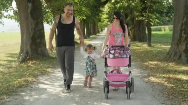 Slow Motion: Neşeli kız güneşli yeşil cadde aşağı iken anne ve babasının ellerini tutar. Kaslı baba ve neşeli anne bahar doğada rahatlatıcı bir yürüyüş sırasında sevimli kızı ile gülümseme.