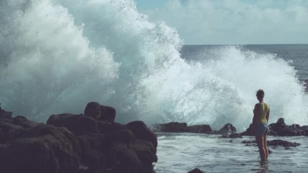 スローモーション 大きな波を見ている若い女性旅行者が激しく岩の海岸に衝突します 泡立つ海水が高く空中に飛び散り イースター島の海岸の黒い岩の上に飛び散る — ストック動画