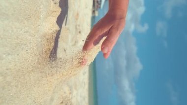 dikey yavaş hareket, yakın çekim: tanınmayan genç kadın sıcak beyaz kum ile güneşli egzotik plajda oynar. Oyuncu kadın turist kum küçük parçacıklar uzak onu nazik elinden sinek sağlar.
