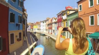Close Up: Tanınmayan genç kadın akıllı telefonu ile renkli bir Venedik caddesinin kanal köprüsünden fotoğraf çekiyor. Çarpıcı Burano pitoresk evleri filme Kadın turist, İtalya.