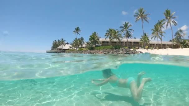 スローモーション ハーフ水中 遠く離れた熱帯の島の近くのガラス張りの海でターコイズビキニダイビングの幸せな女性 さわやかな海の白い砂浜で彼女の休日を楽しむ女性旅行者 — ストック動画