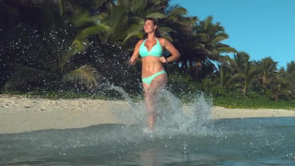慢动作 低角度 欢快的年轻女子在空旷的热带岛屿海滩慢跑时 在空中喷水 运动女孩在绿松石比基尼飞溅玻璃海水 而慢跑 — 图库视频影像