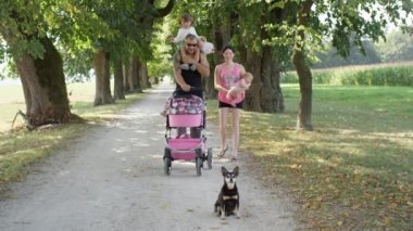 Yavaş Hareket: Neşeli dövmeli anne ve baba iki genç kızı ve küçük köpek ile güneşli bir yürüyüşe çıkmak. Kaslı baba bebek arabasını iterken küçük kızını omuzlarında taşıyor..
