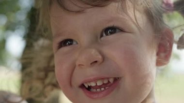 Slow Motion, Close Up, Dof: Gülümseyen küçük kıvırcık saçlı Kafkas kız güneşli ağaç cadde etrafında bakar. Güzel neşeli yürümeye başlayan mesafe içine bakarken ağzına onu küçük parmak koyar