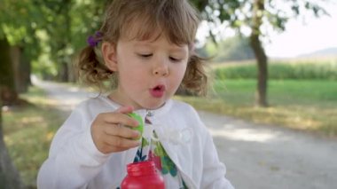 yavaş hareket, portre: oyuncu genç kızı yeşil ağaç caddeye eğlenceli bir aile gezisi sırasında bir sabun köpüğü darbe çalışır. Keyifli küçük kız havada sabun kabarcıkları üfleme, parkta oynar.