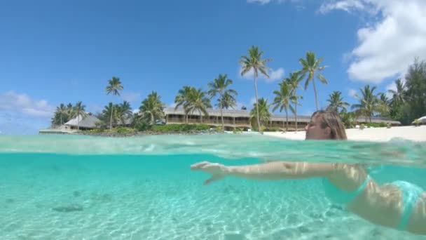SLOW MOTION, HALF UNDERWATER : Joyeux jeune femme en vacances relaxantes nage devant l'hôtel en bord de mer dans les magnifiques îles Cook. Fille souriante en bikini nageant dans l'océan cristallin rafraîchissant .