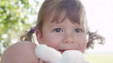 Slow Motion, Portrait, Close Up, Dof: Parktayken oyuncak bebeğiyle oynayan at kuyruklu güzel bir kız. Güneşli bir caddede yürürken oyuncaklarını tutan güzel bir kız çocuğu..