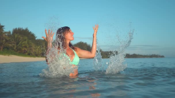 慢动作 复制空间 微笑的白种女人在空中举起双臂 愉快的女孩在绿松石比基尼飞溅在清爽的大海 在她的假期 — 图库视频影像