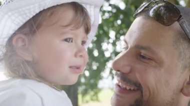Slow Motion, Close Up, Lens Flare: Sevgi dolu baba neşeleniyor ve sevimli masum kızına fısıldıyor. Mutlu baba, sakin yeşil parkta yürürken küçük tatlı kızıyla konuşuyor..