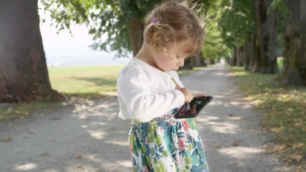 LENT MOTION, FERMER : Adorable jeune fille types avec ses petits doigts sur un téléphone portable moderne. Jolie petite fille jouant à des jeux au parc ensoleillé sur le téléphone portable de son père. Tout-petit tenant un téléphone .