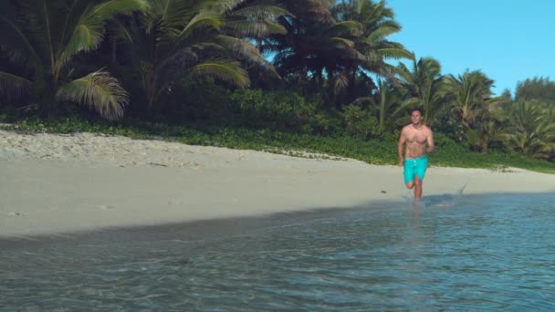スローモーション 素晴らしい夏休みにアクティブな男は 牧歌的な白い砂浜に沿ってジョギング 日当たりの良い熱帯のビーチでリラックスしたランニングを楽しむ若い白人男性 フィット観光客はガラスの水を飛び散らす — ストック動画