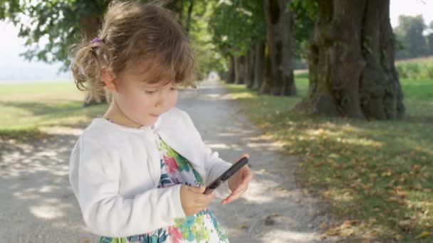 LENT MOTION, FERMER : Bébé fille glisse son petit doigt sur l'écran d'un téléphone cellulaire tout en regardant un dessin animé dans le parc. Belle jeune fille regardant une vidéo drôle sur le téléphone mobile de ses parents .
