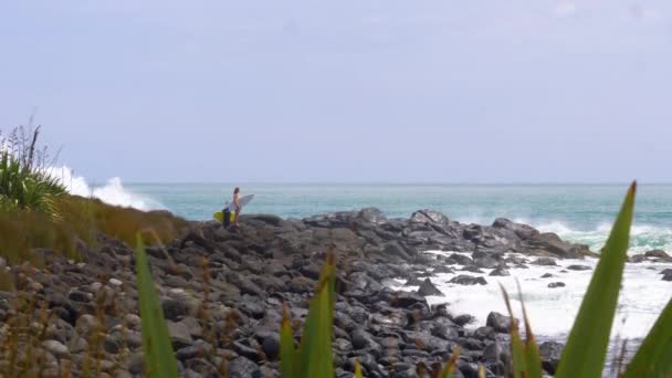 慢动作 复制空间 女人站在她的胳膊下冲浪板 看着海洋 因为巨大的破浪击中岸边 溅过黑色的岩石 强大的海浪在海岸上飞溅 — 图库视频影像