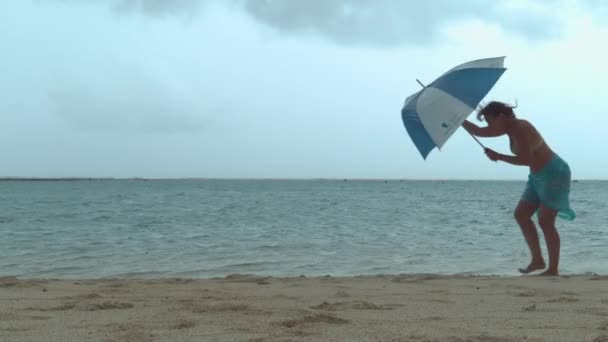 女性の傘は 彼女が砂の熱帯のビーチを歩いている間 強いモンスーン風によって壊れます 若い女性旅行者は 海辺への彼女の旅行中に恐ろしい風の天候に巻き込まれる — ストック動画