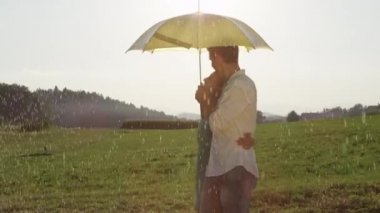 Slow Motion, Close Up, Lens Flare: Aşık neşeli genç çift kırsal taze yaz yağmurzevk. Güzel erkek arkadaşı ve kız arkadaşı yağmurda yürümek ve sarı şemsiye arkasında gizli öpücük.