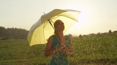 Slow Motion, Close Up, Lens Flare: Neşeli genç kadın bahar yağmurlarında kırsal ortamda dans ediyor. Muhteşem sarışın kız onu sarı şemsiye ile döner ve güzel yaz doğa etrafında bakar.