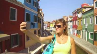 Burano İtalya, Haziran 2017, Gülümseyen genç turist güneşli Venedik, İtalya 'da bir kanal köprüsünde selfie çekiyor. Neşeli kadın kendini Burano 'daki renkli binaların önünde fotoğraflıyor..