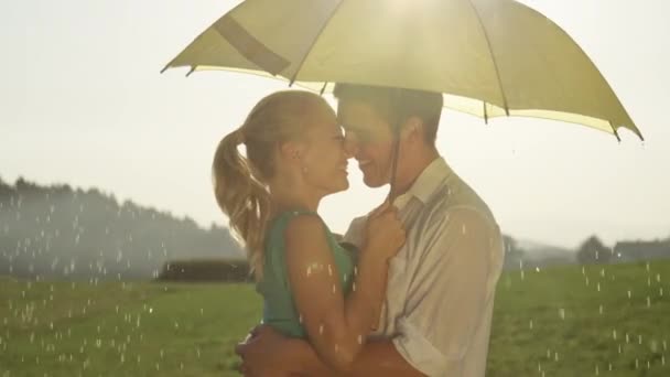 スローモーションクローズアップレンズフレア 楽しいデートに幸せな白人のカップルは黄色の傘の下でキス 熱狂的なボーイフレンドとガールフレンドは 雨の夏の午後に絵のように美しい田園地帯を楽しみます — ストック動画