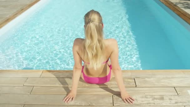 慢动作 无法辨认的年轻女子坐在阳光明媚的泳池边 环顾度假村 运动金发游客在粉红色比基尼浸入她的脚到闪闪发光的水晶清澈的池水 — 图库视频影像