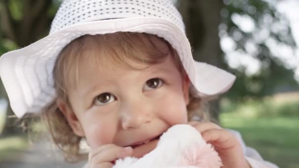慢动作 Dof 可爱的小女儿的肖像与大棕色的眼睛和白色帽子 可爱的小女孩蹒跚学步咀嚼她的玩具 而与她的父母在公园的乐趣 — 图库视频影像