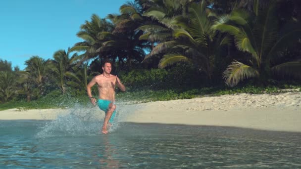スローモーション 幸せな白人男性は エキゾチックな海の浅い部分でシャツなしを実行し 彼の周りの透明な水を飛び散らします 陽気な男は絵のようにヤシの木と白い砂浜に沿ってジョギング — ストック動画