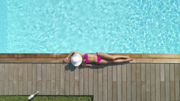 トップダウン シネマグラフ フィットガールは プールで日焼けしながら 彼女の顔の上に帽子を置きます セクシーな若い女性は素晴らしいきらめく豪華なプールでピンクのビキニで日光浴します 女性観光客の寒さ — ストック動画