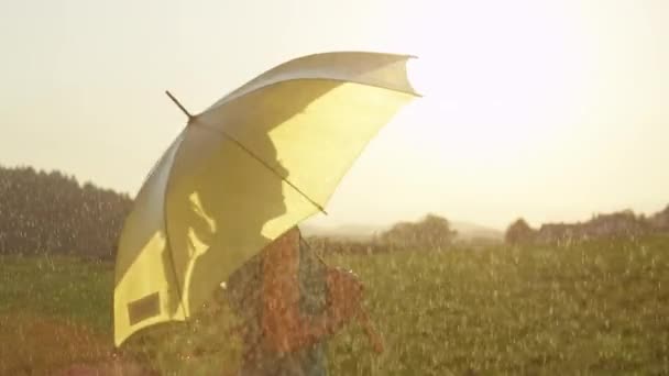 スローモーション クローズアップ サンフレア 幸せな女性は 彼女の黄色の傘を保持しながら 春の雨の中で旋回します ブロンドのポニーテールを持つ気楽な女の子は 雨にもかかわらず 日当たりの良い田舎で笑顔と踊ります — ストック動画