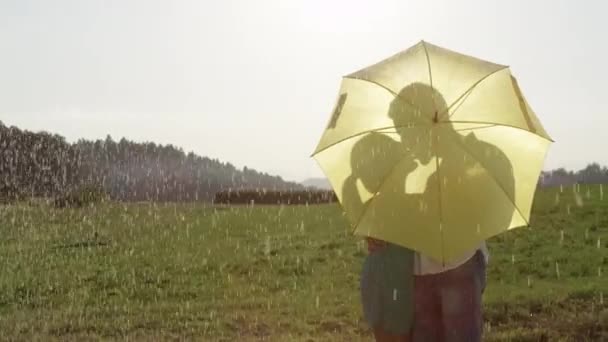 スローモーション クローズアップ ロマンチックなデートに未知の若いカップルは黄色の傘の後ろに隠されたキスです 気楽な少年と女の子は 日当たりの良い田舎で彼らの楽しい散歩中に夏の雨に巻き込まれる — ストック動画
