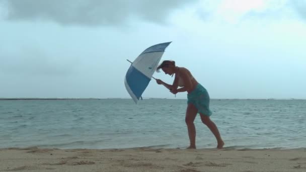 Young female tourist struggles to walk down the sandy beach in the strong monsoon wind. Womans relaxing holiday in Cook Islands is ruined by the miserable weather. Girls umbrella gets destroyed.