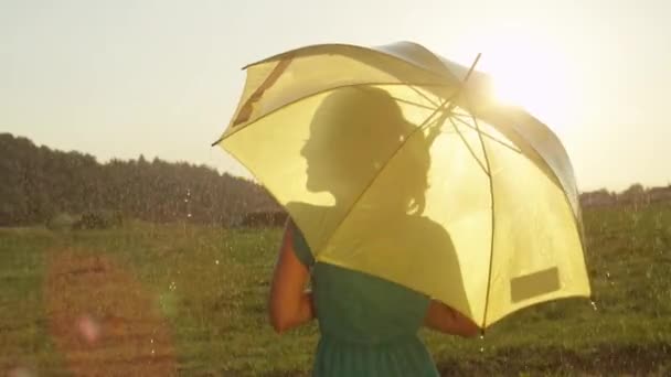慢动作 镜头火焰 迷人的年轻金发女郎拿着黄色伞快乐地看着她的肩膀 同时在风景如画的绿色草地上享受凉爽的夏季降雨 — 图库视频影像