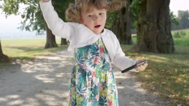 LENT MOTION, FERMER : Petite fille ludique saute autour de l'avenue de l'arbre vide chantant à la musique jouant sur le téléphone mobile de ses parents. Joyeux bébé fille avec des tresses dansant dans le parc ensoleillé 