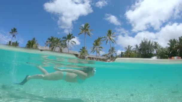 HALF UNDERWATER, SLOW MOTION : Jeune voyageuse en bikini turquoise profitant d'un voyage de plongée en apnée autour d'une île exotique. Femme plongée avec tuba passé superbe hôtel en bord de mer près de la plage de sable blanc .