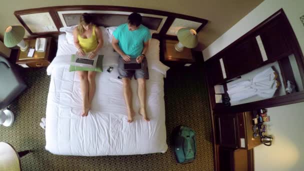 自上而下 年轻的高加索夫妇在越南度假工作在他们的笔记本电脑上 而坐在国王大小的床上 女性和男性游客在舒适的酒店房间上网 — 图库视频影像